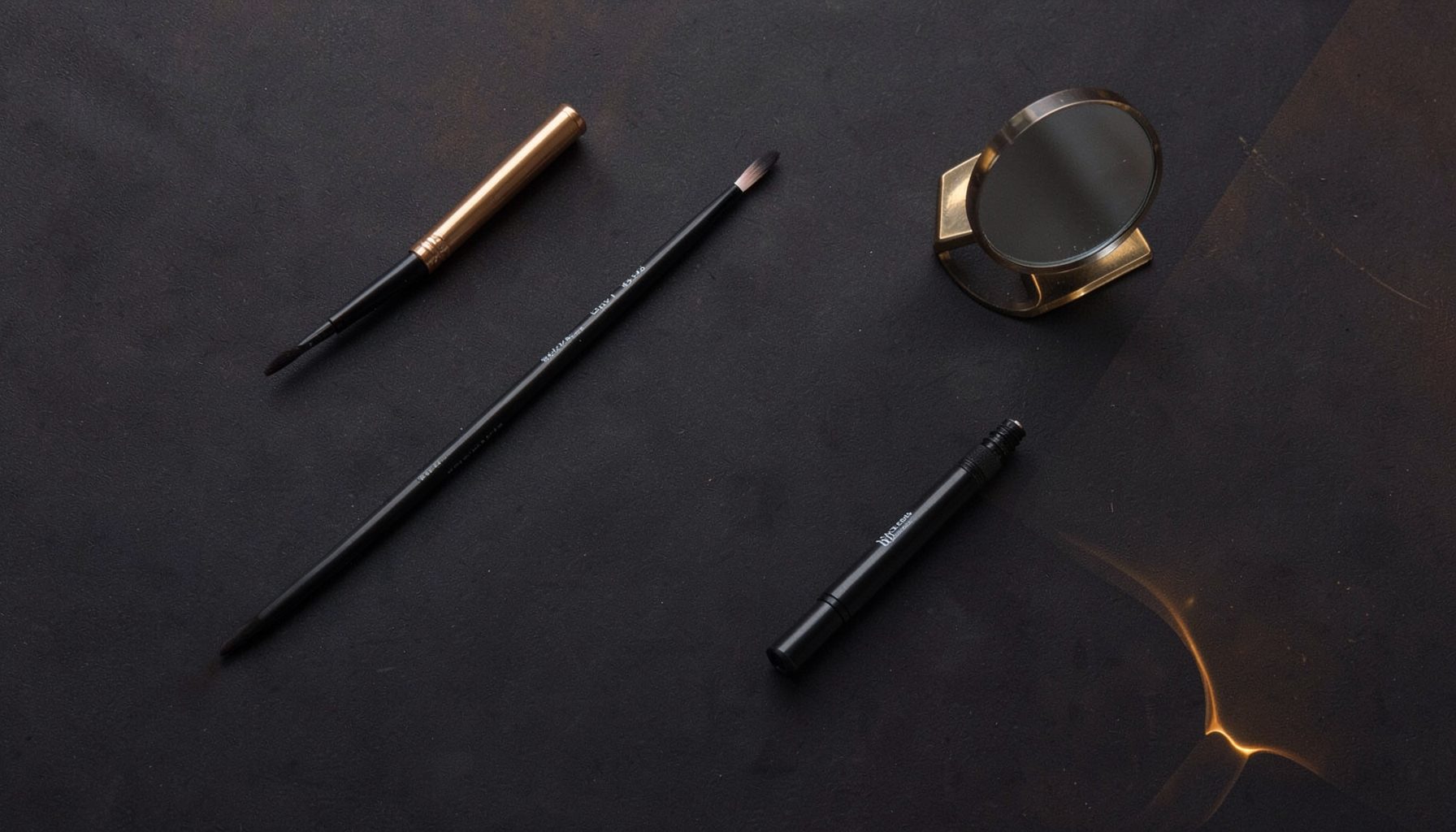 Artistic overhead view of eyeliner tools — a fine-tip brush, dark liner pencil, and small mirror on a deep charcoal surface with warm amber accents