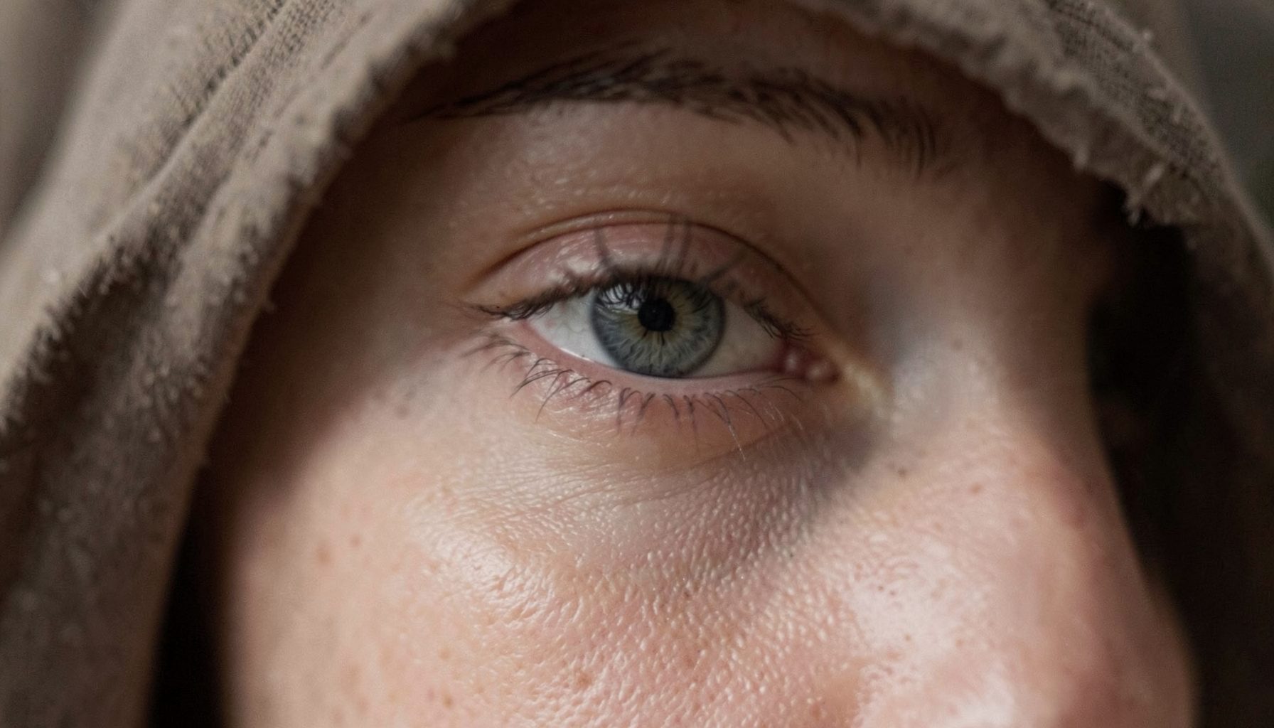 Softly lit eye detail showing a subtle almond shape with partial hooding, neutral earthy tones, understated and natural mood