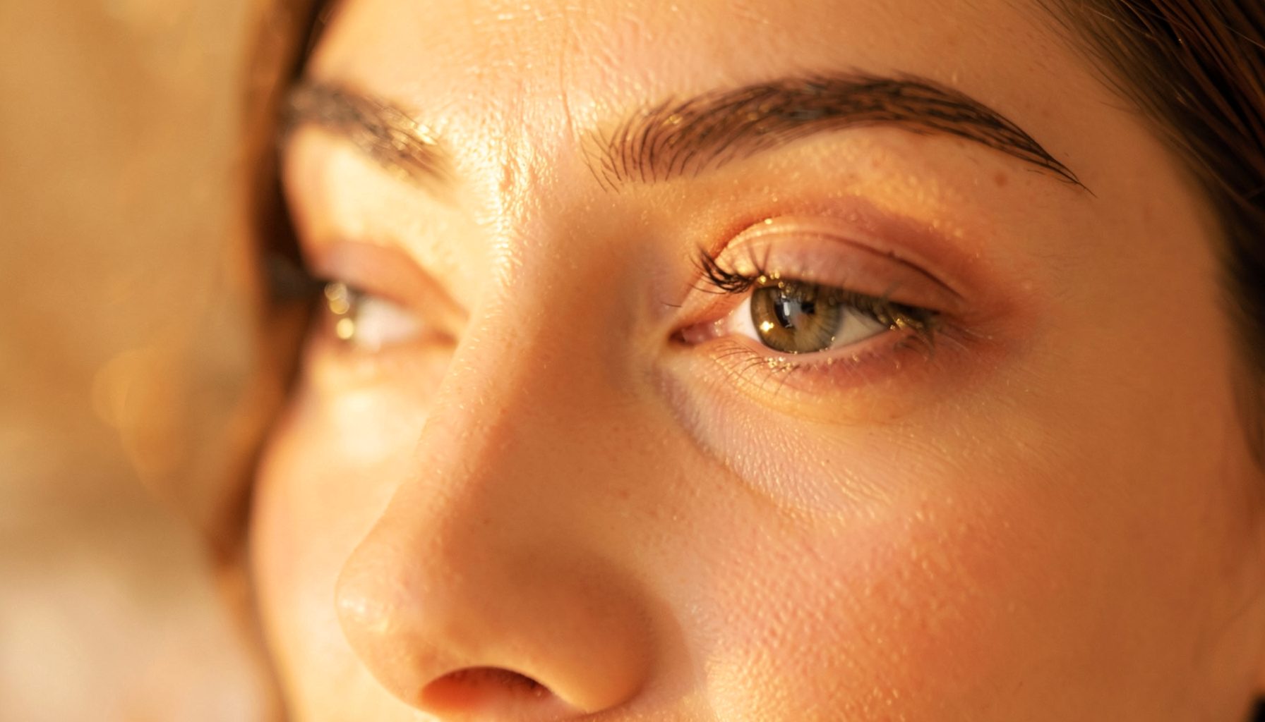 Close-up of a woman's eye area with soft golden light, warm amber tones, serene and elegant mood suggesting a beauty treatment