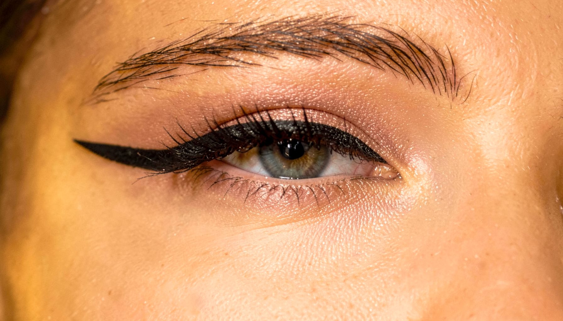 Close-up of a woman's eye with a precise dark winged eyeliner on a warm golden-toned background, elegant and confident mood